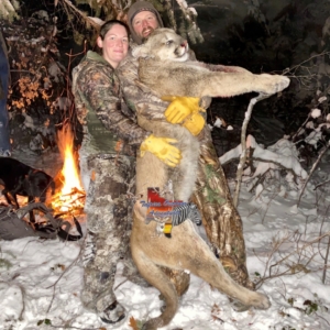 A couple posing in the snow in front of a fire with the large puma they harvested on their Colorado hunt.